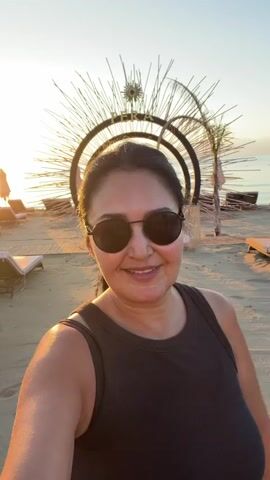 A woman with dark hair and sunglasses smiles at the camera on a sandy beach at sunset. She is wearing a dark tank top. Behind her, there is a large, sunburst-like decoration with a circular frame and palm fronds. Beach chairs are visible in the background. The sun is setting, casting a warm glow over the scene. The woman puckers her lips and then smiles again, as if posing for the camera.