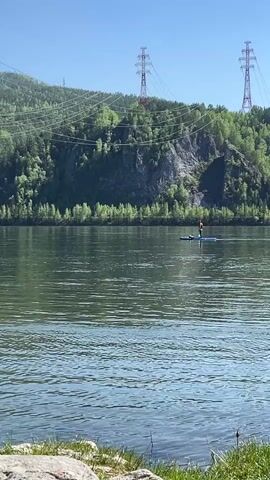 A person is paddleboarding on a calm body of water under a clear blue sky. The paddleboarder, wearing an orange top or life vest, stands on a blue paddleboard in the middle distance. In the background, a lush green forested hillside rises, featuring exposed rocky cliffs. Several large power lines traverse the upper part of the hills, supported by two prominent transmission towers visible against the sky. The foreground shows a partial view of a rocky and grassy shoreline. The time of day appears to be sunny daytime. The scene depicts an outdoor recreational activity in a natural, hilly landscape, consistent with the provided location context of Sliznevo, Russia. No text is visible.