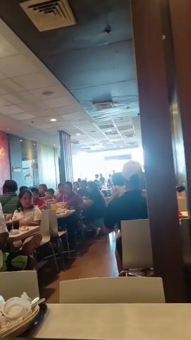 The video appears to be shot in a fast-food restaurant. In the first few frames, people are seen sitting at tables, eating, and interacting. The lighting suggests an indoor environment with fluorescent lighting overhead. The restaurant appears to be moderately busy. In one frame, a child is seen sleeping on a chair, with their legs extended and feet resting on the seat. The child is wearing jeans and a t-shirt. In another frame, a close-up of a child's face is shown, with their eyes closed, indicating they are asleep. The overall impression is a casual dining setting where some patrons are resting.