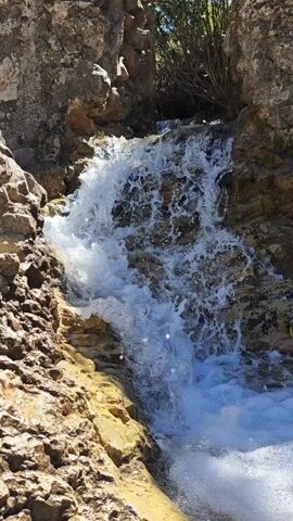 The video shows a small waterfall cascading over rocks. The water is clear and frothy, and the rocks are various shades of brown and tan. There is some greenery visible in the background. The day is sunny.