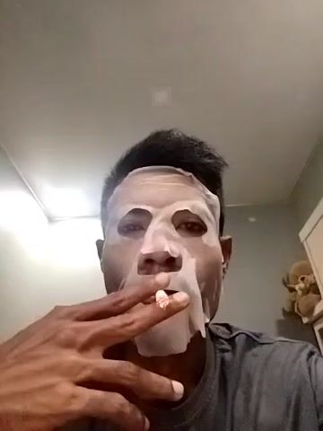 A low-angle shot captures a person wearing a white sheet mask on their face, while holding and smoking a cigarette. The person, appearing to be male with dark hair and a dark complexion, is looking directly at the camera. The sheet mask covers most of their face, including the forehead, cheeks, and chin, with cutouts for the eyes, nose, and mouth.

At the beginning of the video, the person exhales a plume of smoke from the cigarette, which is held between the index and middle fingers of their right hand. They take another drag from the cigarette, which is placed just below the mouth opening of the mask, and then remove it to exhale more smoke. The person is wearing a dark gray t-shirt.

The background is simple, suggesting an indoor setting. A plain, light-colored ceiling with a square light fixture is visible above the person's head. To the right of the frame, a white cabinet or shelf unit is partially visible, with a small brown teddy bear sitting on top. The overall lighting is bright, likely from an artificial source. No specific location cues are visible beyond the general indoor environment. The person's expression is neutral, largely obscured by the mask, with only the eyes clearly visible.