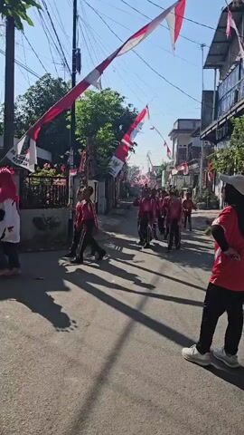 A group of young people in matching red and white uniforms are marching down a narrow street lined with buildings. They are holding flags, and several Indonesian flags are strung across the street overhead, indicating a celebration or event. The sun is shining, casting long shadows on the asphalt road. One person in the foreground is facing away from the camera, also wearing a red top and black pants.