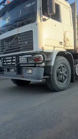 The video shows a large truck with a tarp-covered load, driving down a dusty road. The truck has dual rear wheels and appears to be carrying heavy materials, possibly construction supplies. The side of the truck is visible, showing storage compartments and air tanks. In the background, there are several small structures that appear to be temporary or makeshift buildings, possibly a market or settlement. Some people are gathered near these structures, observing the truck as it passes. The sky is clear and blue, indicating daytime. The overall environment appears to be arid or desert-like.