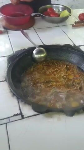 The video shows a close-up of a large pot of food being cooked, likely plov given the setting. A large ladle rests in the pot, which is filled with rice, meat, and vegetables, all simmering in a rich broth. Steam rises from the pot, indicating it's hot and actively cooking.

In the background, a person, appearing to be an older man wearing a traditional skullcap (doppi), is shown preparing tomatoes. He holds a tomato in his left hand and is using a knife in his right to peel it. A red bowl sits on a wooden cutting board in front of him, and a larger silver bowl contains more tomatoes and some sliced green peppers. Another red tomato is visible on the table to the right of the silver bowl, near a set of knives.

The scene appears to be outdoors or in a semi-outdoor kitchen, with a tiled countertop and a glimpse of greenery and what looks like a light-colored roof or awning in the distance. The lighting suggests it's daytime. The overall atmosphere is one of food preparation in a domestic or communal setting, likely in Uzbekistan, as hinted by the man's attire and the dish being prepared. There is no visible text in the media.