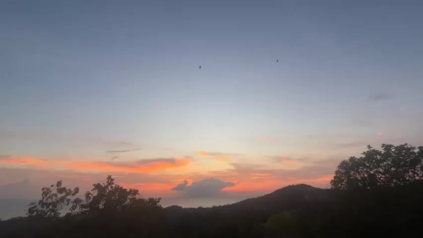 The video shows a wide shot of a landscape at sunset. The sky transitions from blue at the top to orange and pink near the horizon, where the sun is setting. A few scattered clouds are visible, tinged with orange. Silhouetted mountains and trees form the lower part of the frame. Two birds are flying in the upper part of the sky.