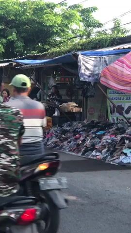 A person wearing a mask and a gray jacket is sitting on a black motorcycle with green accents in a street market. The motorcycle is facing left. Behind the motorcycle, there is a stall with a blue tarp roof. Several pairs of shoes are displayed on the ground in front of the stall. To the left of the motorcycle, another person wearing a floral dress is standing near a pile of goods. There are trees and buildings in the background. The overall scene appears to be a busy outdoor market.