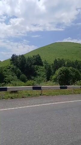 The video shows a roadside view with a large, green, grassy hill in the background. The sky is partly cloudy. In the foreground, there is a road with a guardrail. Various objects are visible along the roadside, including a silver car, a trailer that appears to be a mobile beehive, a chair, and some signage. A dark-colored SUV drives past in the foreground towards the end of the clip. The overall scene suggests a rural or mountainous environment during the daytime.
