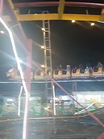 The video shows a spinning carnival ride at night. The ride is shaped like a caterpillar or a centipede, and it is full of passengers. The ride is brightly lit with white and colored lights. The camera follows the ride as it spins around, providing a close-up view of the colorful cars and the riders.