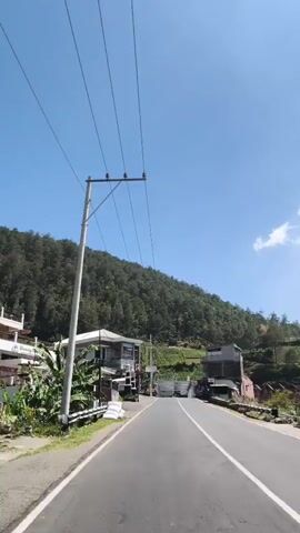 A person rides a scooter down a paved road. The road is flanked by buildings and lush green trees on a mountainside. Power lines stretch across the sky, which is a clear blue with a few white clouds. The scooter is traveling away from the camera.