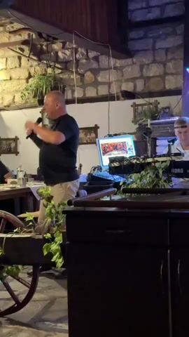 A bald man in a black t-shirt and khaki pants stands on the left, holding a microphone and speaking. He is positioned near a decorative cart with green vines. Behind him, to the right, a man in a white shirt plays a keyboard. The environment appears to be an indoor venue with stone walls, wooden beams, and plants. There is a computer monitor displaying an image, along with various musical equipment. The lighting is dim, with some purple and green hues.