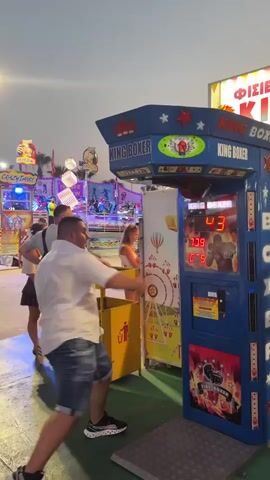 A man in a white short-sleeved shirt and denim shorts plays a boxing arcade game in an outdoor amusement park setting. The game machine is blue with "KING BOXER" written on it and displays a digital score. The man has just thrown a punch, and the score is registering 623. In the background, other amusement park rides and structures are visible, including a Ferris wheel. Two women and another man are standing near the game. The sky is a light blue, indicating it is daytime or early evening.