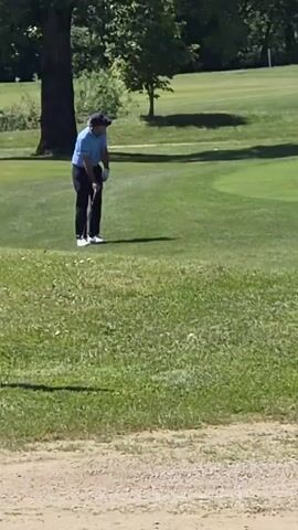 A man in a blue shirt, dark pants, a hat, and white shoes is walking on a golf course. He is holding a golf club in his right hand and is walking toward a green. The camera pans to show a sign that says "Golf Course Area" with a symbol indicating no golf carts. The camera pans back to show the man on the golf course. He disappears into some tall grass.