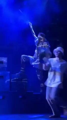 The video shows a stage performance lit with blue lights. Two performers are visible, dressed in white and silver clothing. One performer is standing on a stack of black boxes, holding a long, thin object, possibly a microphone or prop. The other performer is moving around the stage, at one point holding up a shiny, spherical object. The performers appear to be engaging in a theatrical or musical performance. The stage is dark with blue lighting.