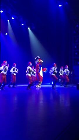 A group of approximately ten individuals, dressed in pirate costumes consisting of white shirts, red and white striped pants, and red headscarves, perform on a stage alongside a large red, yellow, and blue parrot mascot. They are all dancing in unison, lifting their knees and moving their arms. The stage is dimly lit with strong blue lighting, including spotlights on the performers. A person in dark clothing is partially visible on the right side of the stage, near the back.
