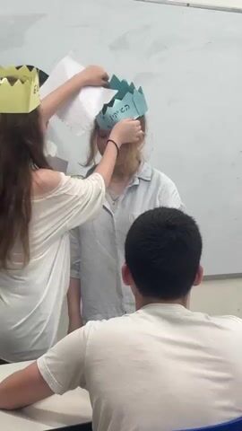 In a classroom setting, several young people are participating in an activity involving paper crowns. A young woman in the center is wearing a light blue paper crown on her head, which has some Hebrew writing on it. Another young woman, with long brown hair, is adjusting the crown on the central figure's head. She is wearing a white off-the-shoulder top and a yellow paper crown with a floral design on her own head. A boy with dark hair is seated in the foreground, facing away from the camera, wearing a white t-shirt. Another boy is seated beside him, also facing away from the camera. Behind the central figure, another young person with dark hair is visible, wearing a white t-shirt. The room appears to be a classroom with a whiteboard on the left and a bookshelf on the right. The lighting suggests it is daytime. The overall atmosphere appears to be one of engagement and playful activity.