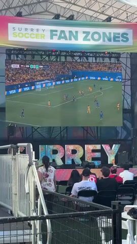 A large outdoor screen displays a soccer match. The score is tied at 0-0 in the 59th minute. On the screen, a player in a blue uniform is dribbling the ball towards the goal defended by a player in yellow. Another player in blue is positioned near the goal. The stadium seats are filled with spectators wearing yellow clothing, suggesting they are fans of one of the teams. Below the large screen, a banner reads "SURREY FAN ZONES." In the foreground, a few people are seated on chairs, watching the game on the screen. A woman in a floral shirt is standing and looking towards the screen. The environment appears to be an outdoor event space with barriers and a metal structure visible.
