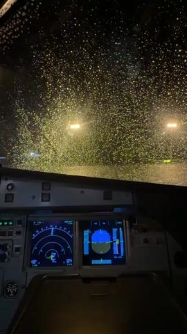 The video provides a view from the cockpit of an aircraft, looking out through the windshield at night. The windshield is covered in numerous water droplets, suggesting it is raining. Distant yellow lights are visible through the rain, possibly airport runway lights or streetlights.

The focus of the foreground is the aircraft's instrument panel. Two main displays are visible. The left display shows a radar-like screen with circular arcs and a crosshair indicating a target or waypoint. Several numbers and indicators are present on this screen, including "2.8 KM" and "VFR IFR", suggesting flight path and navigation information.

The right display is a more complex artificial horizon or flight mode display. It shows a blue and black horizon line, indicating the aircraft's pitch and roll. Vertical bars on the right side likely represent altitude or speed, with numerical values like "17500" and "25000". Other indicators and labels such as "CLB", "NAV", and "FCS" are also present.

The overall atmosphere conveyed is one of a potentially challenging flight condition due to the rain and darkness, but the illuminated and functional cockpit displays suggest the pilots are in control. The time of day is clearly night. While no people are explicitly visible, the perspective implies the presence of pilots or crew. The location context of San Juan Bautista, Peru, doesn't provide specific visual cues within the frame itself.