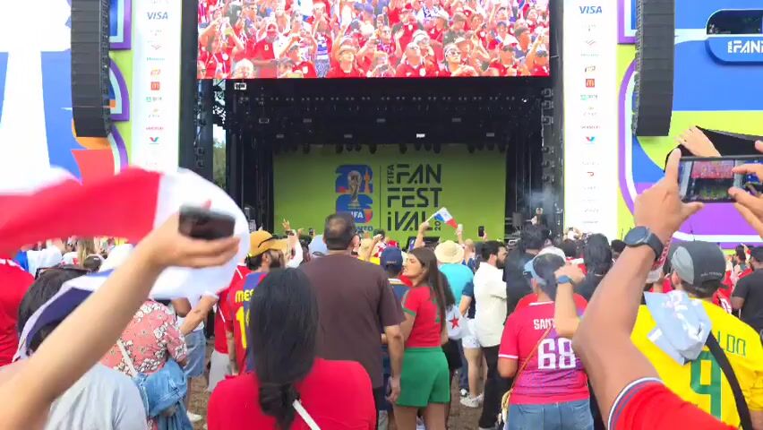 The video captures a lively outdoor event, likely a FIFA Fan Fest, given the large screen displaying the event logo "FIFA FAN FEST PANAMA 2026". A large crowd of people, many wearing red and white, are gathered in front of a stage. The crowd appears excited, with many holding up phones to record or take pictures. Some individuals are waving flags, one of which appears to be the Panamanian flag. The atmosphere is festive, suggesting a viewing party or a concert. The screen above the stage also shows a large group of people, presumably from a past event or a live broadcast, also appearing to be in celebration mode. The overall scene is one of a public gathering with a focus on entertainment and sports.