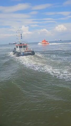The video captures a nautical scene from the perspective of a moving vessel. In the foreground, the choppy, green water churns, leaving a white wake. Two boats are visible in the middle ground: a smaller, white and black tugboat in the left foreground, moving away from the camera, and a larger, orange and white rescue boat further out in the water, also moving away. In the background, several other ships are present on the horizon. The sky is blue with scattered white clouds.
