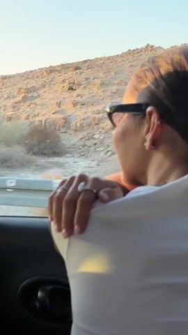 A medium shot captures a person's back and shoulder as they look out of a car window. The person is wearing a white shirt and black-rimmed sunglasses. A ring is visible on their left hand, which rests on their shoulder. The environment outside the car appears to be a desert landscape with rocky terrain and sparse vegetation. A black and white striped pole is visible on the left side of the frame. The lighting suggests it is either sunrise or sunset, casting a warm glow on the scene.