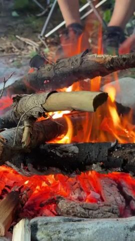 The video shows a close-up view of a campfire with several logs burning. The flames are orange and yellow, and the embers are red. In the background, two people are sitting in camping chairs. One person's legs and feet are visible, wearing dark pants and shoes. The other person's legs and feet are partially visible. The environment appears to be outdoors, with dirt and rocks visible on the ground. The overall scene depicts a camping or outdoor recreational setting with a focus on the burning fire.