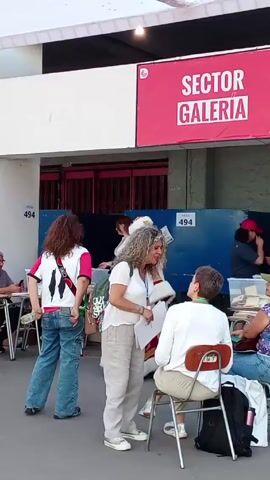 The video shows the exterior of a building with a sign that says "SECTOR GALERIA" in large white letters on a red background. Several people are standing and sitting around tables, likely election workers. A woman with curly hair stands and talks to a person sitting on a chair. A person dressed as Santa Claus approaches and speaks to the seated individual. There are tables with papers and election materials, and signs indicating "494."