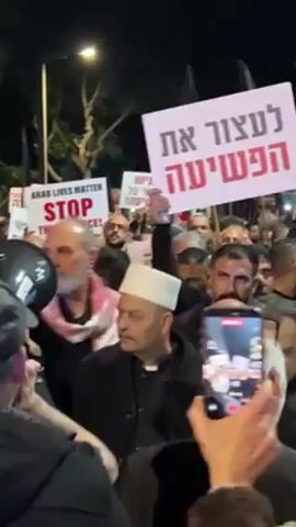 A crowd of people are gathered at night. Some are holding signs; one says “Arab lives matter, Stop the Violence!” in English. Another sign held by a man in the front reads in Hebrew. One man near the front is wearing a white cap and black clergyman's robes. People are chanting. Someone is recording the scene with their phone. The crowd appears dense and illuminated by streetlights and device screens.