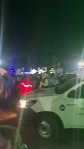 The video depicts a bustling nighttime scene featuring a dense crowd gathered around a white pickup truck marked with "Hospital Villa La Angostura" and emergency vehicle lights. The crowd includes individuals of various ages, with a child seen perched on someone's shoulders. A person in a bright red jacket with reflective stripes stands out in the foreground. The atmosphere is lively, possibly celebratory, against a backdrop of tents and lighting.