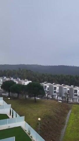 The video shows an outdoor view of a neighborhood with modern white buildings.  The sky transitions from overcast to partly cloudy. There are hills covered with trees in the background. There is a large grassy area in the foreground.  A white van is parked near the buildings. Later, the camera angle shifts to show more of the town beyond the green space. The view then focuses on the row of modern buildings, highlighting their balconies.