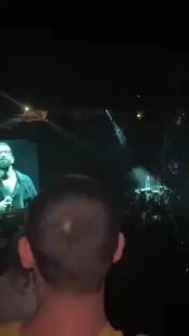Here is a description of the video.
The video shows a concert with a male singer on stage. There's a large screen displaying a man's face to the left. The concert has a crowd of people, and there are several lighting structures above the stage. The stage itself is illuminated with green-tinged light and smoke. The video is shot from the perspective of someone in the audience. The head of the person recording the video is visible in the foreground.