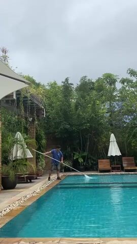 A person wearing a blue t-shirt and shorts is standing on the edge of a swimming pool, holding a long pole with a net attached. They are using the net to scoop debris from the water. The pool is surrounded by a stone walkway and lush green vegetation. There are lounge chairs and umbrellas visible in the background, suggesting a resort or hotel setting. The sky is overcast.