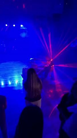 The video shows a crowded event with people dancing. The room is dark and filled with blue and red lights, possibly from a DJ setup or stage lighting. Lasers are visible, creating beams of light that crisscross the space. Some individuals are holding up phones, likely recording the event. The attendees appear to be dressed in formal or semi-formal attire.
