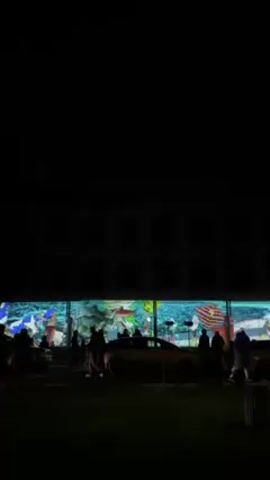 A night scene shows an illuminated building with people standing in front of it, along with cars. A display projected onto the building depicts a chaotic mix of images including an American flag. As the video pans right, it reveals a light pole with a red light on top and a green light below. People are walking around in the foreground. The environment appears to be an outdoor public space.