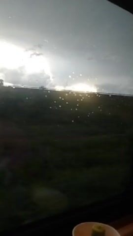 The video shows a view through a window, possibly from a vehicle, with water droplets on the glass. The background is a landscape with a bright, overcast sky. A road or elevated structure is visible in the distance with a truck on it. The foreground is dark and blurry. There is a small cup with something green in it in the bottom right corner.