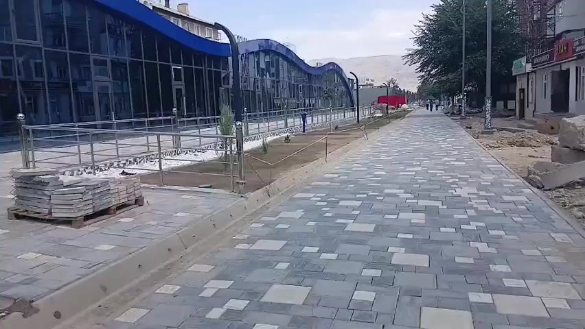The video pans across a newly paved sidewalk next to a modern building with a wavy blue roof and a glass facade. Piles of paving stones are stacked on a pallet on the left side of the sidewalk. A metal railing runs along the edge of the building. The building appears to be under construction or recently completed, with scaffolding visible in the background and some unfinished areas. There are a few small trees and plants planted along the edge of the sidewalk. In the distance, people are walking along the sidewalk, and there are some buildings and hills visible. The sky is cloudy.