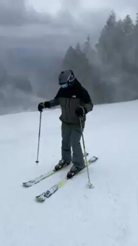 A person in full ski gear stands on a snowy hill. In the background, a snow machine is actively blowing snow, creating a foggy mist. The person is wearing a helmet, goggles, a black and grey jacket, and grey pants. They are holding ski poles and standing on skis with bright green accents. The environment is overcast with a line of evergreen trees in the distance. The person adjusts their gloves before the video ends.