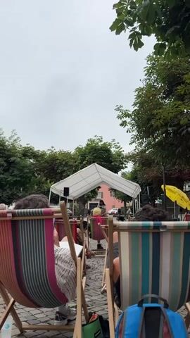 The video shows an outdoor gathering of people seated in lounge chairs, facing away from the camera. The chairs are striped in various colors, including pink, orange, green, and white. In the background, there is a white tent structure, possibly a stage, with a person standing in front of it. To the right of the tent, a bright yellow umbrella is visible. The ground is paved with cobblestones. Green trees with dense foliage are present on both sides of the scene, and the sky is overcast with a cloudy atmosphere. Several backpacks and bags are visible near the chairs. The focus is on the backs of the seated individuals and the setup for an outdoor event.