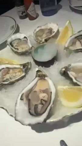 A plate of raw oysters on crushed ice sits on a white table. There are six oysters in their shells arranged around a small, mint-colored bowl. Lemon wedges are nestled between the oysters. Steam emanates from the mint-colored bowl. Tabasco sauce and a pepper grinder are in the background, along with a partially visible white plate and a blue water bottle. A hand briefly enters the frame, wafting away the steam from the bowl.