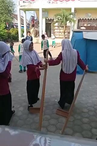 Four young girls wearing hijabs and long skirts stand in a courtyard. Two of the girls in the foreground are holding tall wooden stilts, one in each hand, preparing to walk on them. One girl in a pink shirt and white hijab faces the camera, holding the stilts. Another girl in a similar outfit is to her right, also holding stilts. Two other girls are partially visible to the left, also in pink shirts and white hijabs. In the background, other children are seen playing and walking. The ground is paved with hexagonal tiles. A building with a red and white banner is visible behind the children.