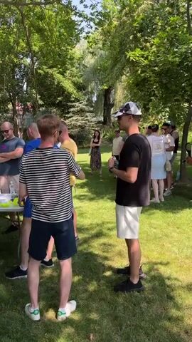 This is a medium shot video, captured outdoors in a park-like setting with lush green trees and grass, on a sunny day. The video is composed of multiple short clips, with the camera panning and zooming to provide different perspectives of a gathering of people around two white folding tables. In the foreground, several individuals are visible, some interacting with each other, others observing. The people are casually dressed, suggesting a relaxed social event, possibly a picnic or a party. Objects on the tables include various small items such as toys, balls, rings, and what appear to be small prizes or game components. Some people are standing, others are leaning towards the tables, indicating engagement with the activities or displays. The background shows more people mingling and trees providing shade, creating a pleasant outdoor atmosphere. The camera movement suggests a dynamic recording of the event, capturing moments of interaction and general scene overview.