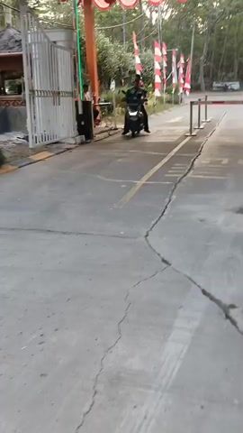 A person on a motorcycle exits a gate and passes by a security guard. The motorcycle is black and the rider is wearing a green shirt. The security guard is wearing a yellow vest and black pants. There are red and white flags displayed on poles in the background, as well as trees. The ground is asphalt and cracked. There are two barriers that are partially lowered. The video is shaky and appears to be taken from a low angle.
