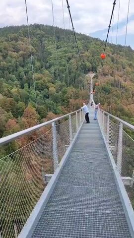 A first-person perspective video shows a long suspension bridge with a metal grate walkway and metal railings on either side. Two people are visible at the far end of the bridge, one in a white shirt and blue pants, the other in a blue shirt and darker pants. They are standing and appear to be looking out at the scenery. The bridge is suspended over a deep valley filled with dense green trees that cover the rolling hills. Several orange spherical objects are visible hanging from wires above the bridge. The sky is cloudy. The camera pans slightly to the right, revealing more of the mountainous, forested landscape and a small village in the distance. A voice in the background says, "We were already at one post, right? Well, it's not here."