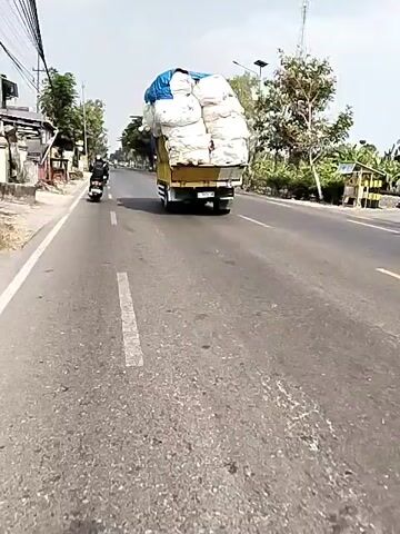 This is a first-person perspective video taken from a moving vehicle, likely a motorcycle, on a paved road. The camera is pointed forward, showing the road ahead and the surrounding environment. In the foreground, the asphalt road surface is visible with a white dashed line marking the lane. To the left of the road, there is a sidewalk area with some vegetation, a fence, and buildings that appear to be shops or houses. A motorcycle is seen in the distance on the left side of the road. The main focus of the video is a large yellow truck driving in the same direction as the camera. The truck is heavily overloaded with large, white, irregularly shaped bags, covered partially by a blue tarp. The load is piled high and appears to extend significantly above the truck's bed, creating a very tall and precarious-looking cargo. The truck is in the right lane of the road. Beyond the truck, the road continues into the distance, flanked by trees and vegetation on both sides. The sky is visible, appearing bright and possibly overcast. The overall impression is of a typical roadside scene with a heavily laden truck driving on a sunny day.