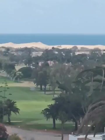 The video shows an outdoor daytime shot of a coastal golf course landscape. In the background, a sandy beach stretches along the blue sea under a clear sky. The foreground features a golf course with green fairways, interspersed with various trees including palm trees. There are white buildings with red-tiled roofs scattered throughout the area. A small pond reflects the sky amidst the trees. The camera is relatively still, providing a wide view of the scene.
