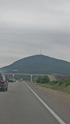 A video shows a wide, paved road with a white line marking the lanes. The camera is in a moving vehicle, and the perspective is from the driver's side. In the foreground, a dark SUV with a roof carrier is visible, driving in the same direction as the camera. Other vehicles are also on the road. In the background, a large, rounded mountain covered in dense green trees dominates the landscape. A radio tower is situated at the very peak of the mountain. A bridge spans across the road in the middle ground, leading towards the mountain. The sky is overcast and a dull gray. The environment appears to be rural, with grassy areas and scattered trees lining the roadside.