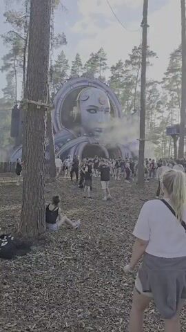 The video shows a crowd of people gathered in a wooded area in front of a large, elaborate stage structure resembling a giant robotic head. The head has glowing purple lights and appears to be emitting smoke or fog. People are casually dressed and milling around, some sitting on the ground. The overall atmosphere suggests an outdoor event, possibly a festival or concert.