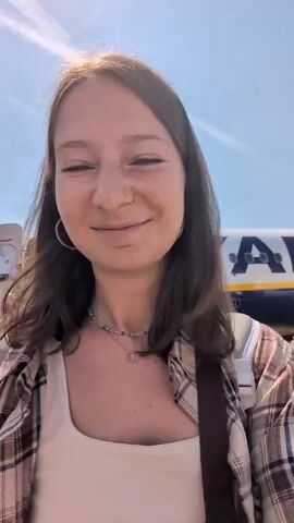 A young woman with brown hair and freckles smiles at the camera. She is wearing a plaid shirt over a cream-colored tank top and a beaded necklace. Behind her, a Ryanair airplane is parked on the tarmac. The sun is shining brightly, and the sky is blue. The woman appears to be at an airport, possibly about to board a flight.