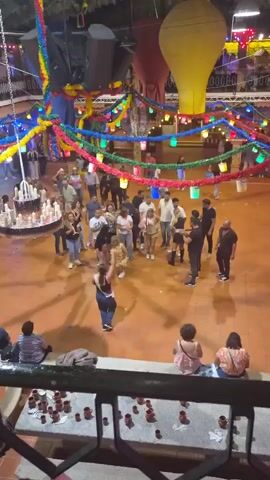 This is a high-angle, real-time video showing an outdoor gathering of people. Colorful streamers and lanterns are strung across the scene, suggesting a festive atmosphere. In the center of the frame, a group of people is gathered in a circle, with some individuals dancing and interacting. A baby is being held by one of the individuals in the circle. Other people are standing around the perimeter of the circle, observing the activity. In the foreground, a railing is visible with several small terracotta-colored cups arranged on it. The ground is paved with orange-toned tiles. The lighting suggests it is either evening or dusk, with artificial lights illuminating the area. Some people in the background are visible, also engaged in activities or observing.