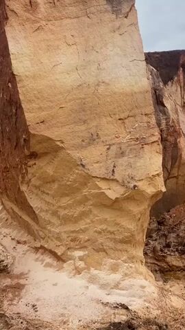 The video pans up a vertical cliff face. The cliff is made of layered sediment, with the top layer being a dark reddish-brown soil with some grass growing out of it. Below this is a lighter tan colored layer that shows distinct horizontal striations. The bottom of the visible cliff face is a lighter sandy color. The overall impression is of an eroded geological formation.