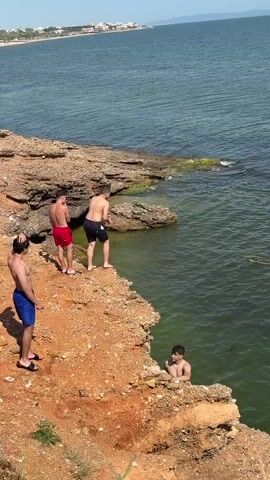 The video captures a scenic coastal setting on a sunny day, featuring several young men engaging in cliff jumping. The scene is dominated by a reddish-brown, rocky cliff that overlooks a calm, blue-green sea.

In the initial shot, one man, wearing dark shorts, is seen mid-air as he jumps from the cliff edge into the water below, creating a significant splash upon impact. Another man is already swimming in the sea nearby. Two more men are visible on the cliff, observing or preparing to jump; one in red shorts and another in blue shorts. In the distance, across the expanse of the sea, faint outlines of buildings or land can be seen under a bright, partly cloudy sky.

The video then transitions to a broader view of the tranquil sea, stretching out to the horizon under a clear blue sky with minimal cloud cover, emphasizing the vastness of the water. The overall atmosphere suggests a recreational and adventurous activity taking place on a pleasant day.